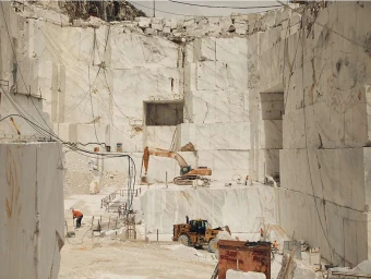 Wide view of a massive open-pit marble quarry with large blocks, heavy machinery, and distant workers by Dave Imms