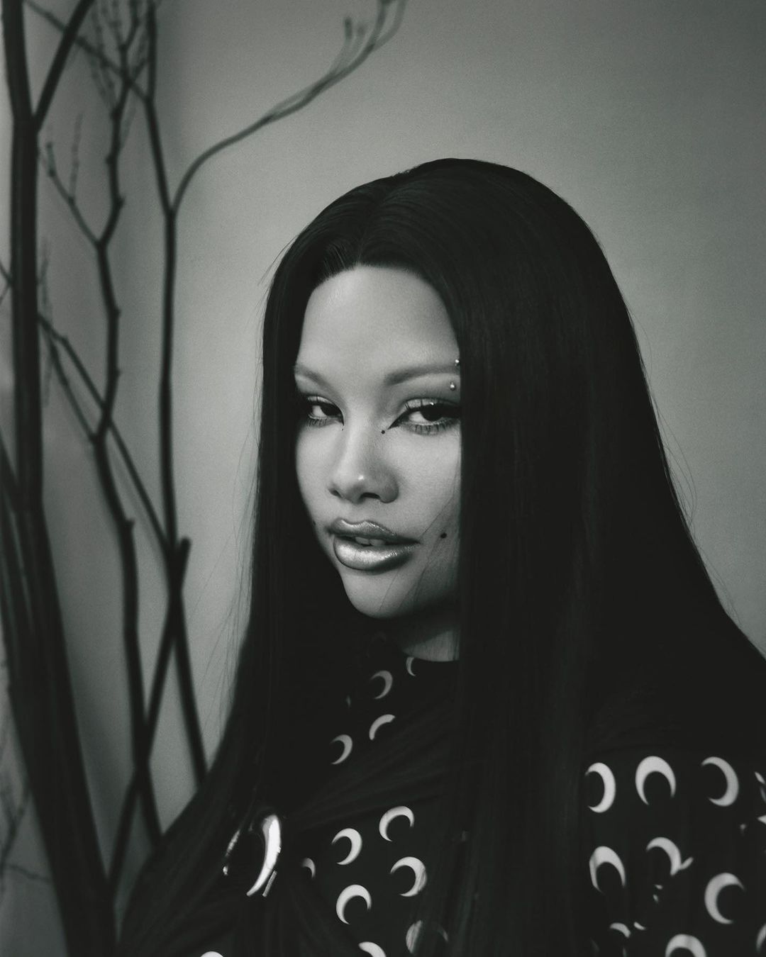 Close-up portrait of a young adult woman with long black hair, glossy makeup, and an ornamental piercing, set against a neutral background.