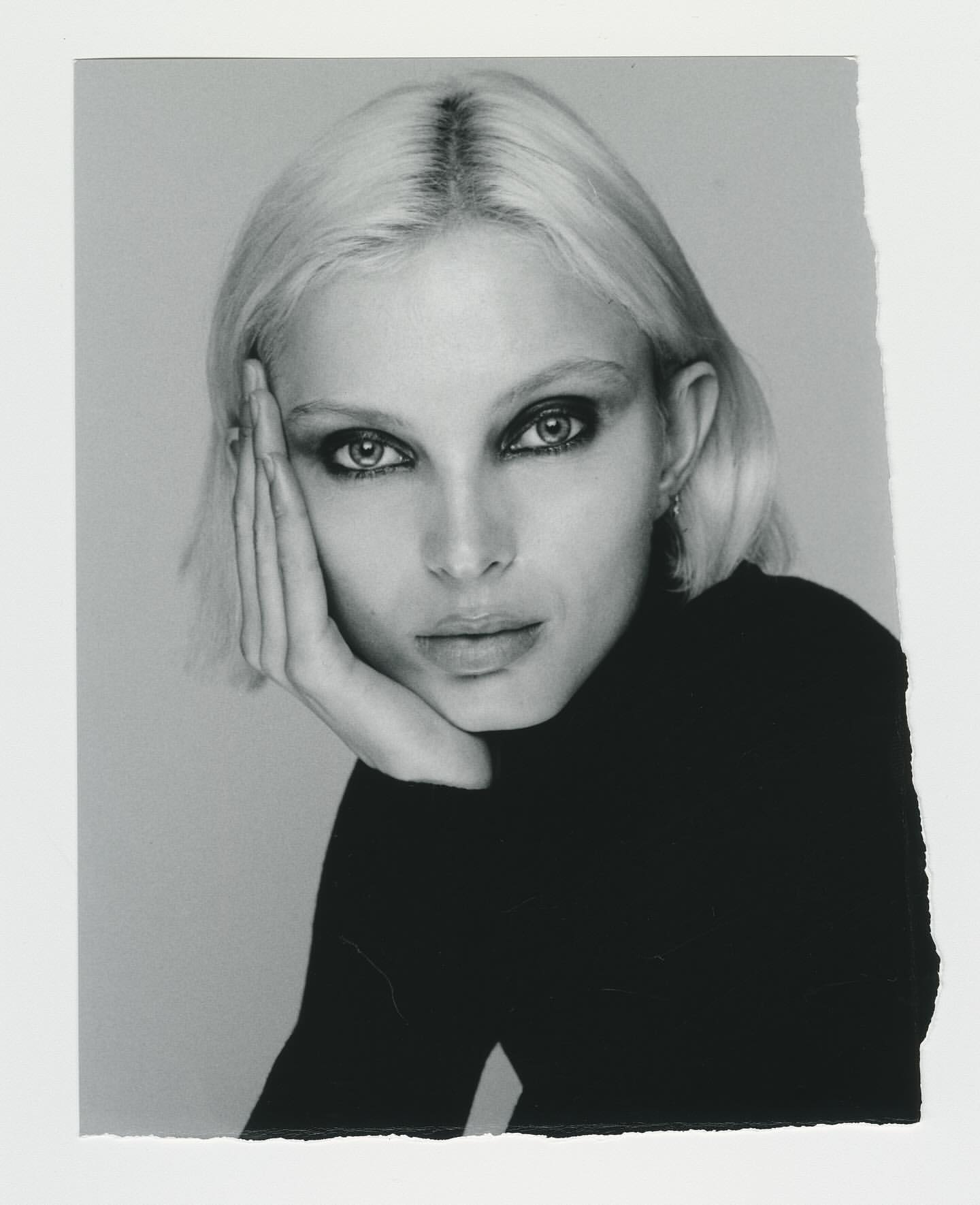 Monochrome portrait of a fair-skinned woman with platinum blonde hair in a black turtleneck sweater, gazing into the camera.
