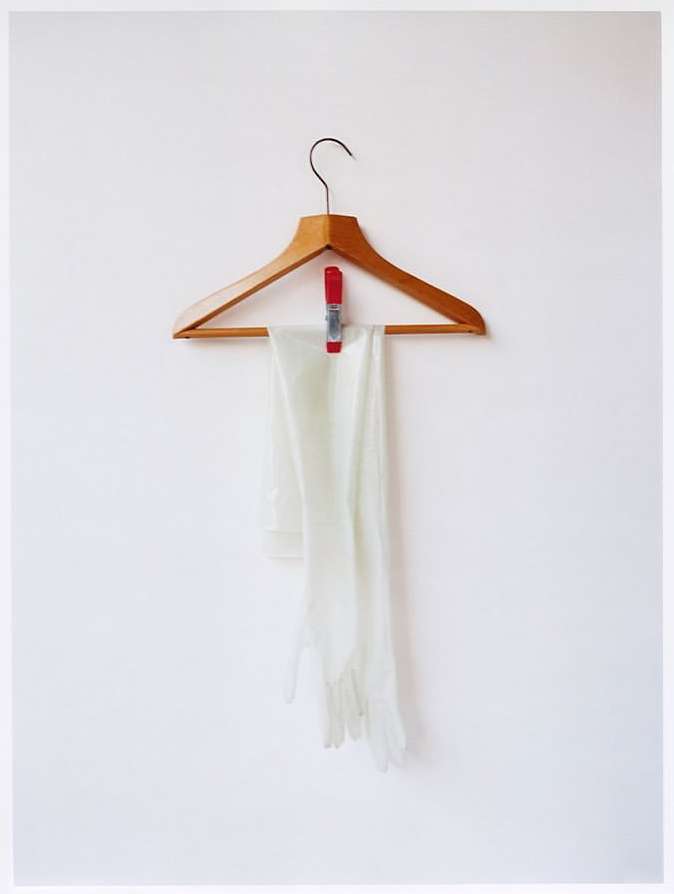 White gloves hang on a wooden hanger against a white wall, held by a red clothespin, providing a simple and elegant composition.