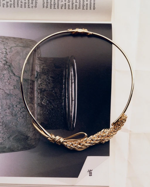 Golden neck ring jewellery with woven metal embellishment laying on the surface of a magazine page. Elegant fashion accessory.