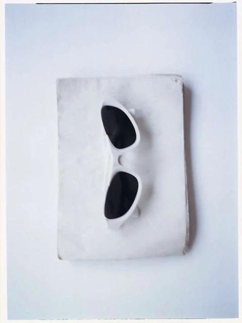 A pair of white-framed sunglasses are positioned on top of a rectangular, stained piece of paper, all set against a white backdrop.
