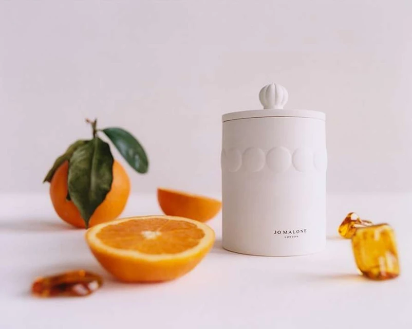 Still life photograph of a Jo Malone candle, orange slices, leaves, and amber stones on a white surface, promoting home fragrance.