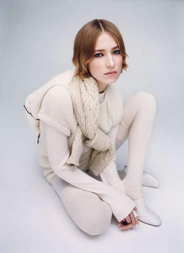 A young woman in an all-white knitwear ensemble poses in a studio setting, with a shallow depth of field and neutral color palette.