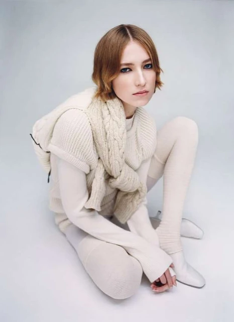 A young woman in an all-white knitwear ensemble poses in a studio setting, with a shallow depth of field and neutral color palette.