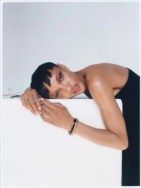 Adult female model with short dark hair in black top poses with bracelets in a light, neutral indoor studio setting.