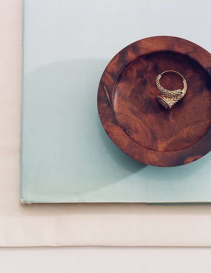 Overhead studio shot of a gold-toned hoop earring with intricate detail set inside a dark wooden bowl, against a two-tone blue and white backdrop.