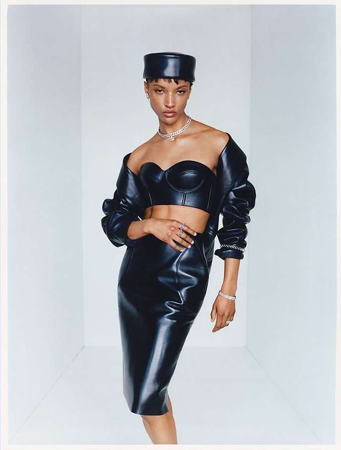 Fashionable young woman in a shiny black two-piece outfit with matching hat posing in a minimalist white room. Jewellery accents.