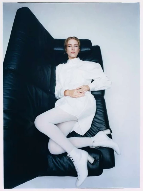 A fair-skinned woman in a white dress and white tights poses on a black leather couch in a photo studio with a white background.