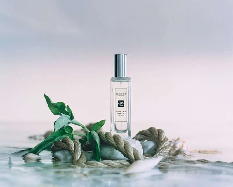 Jo Malone Wood Sage & Sea Salt cologne bottle sitting on rocks and rope in shallow water against a soft blue and pink sky.