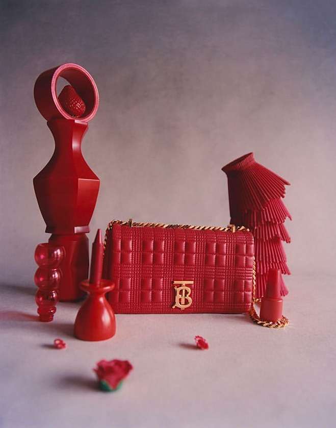 Red Burberry Lola bag with gold chain on a gray surface surrounded by red design objects, creating a visually striking composition.