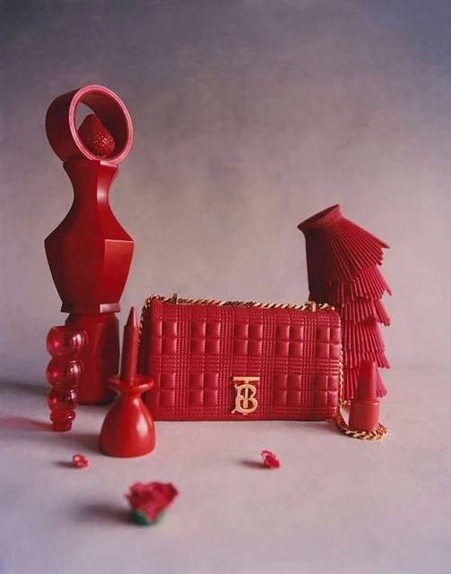 Red Burberry Lola bag with gold chain on a gray surface surrounded by red design objects, creating a visually striking composition.