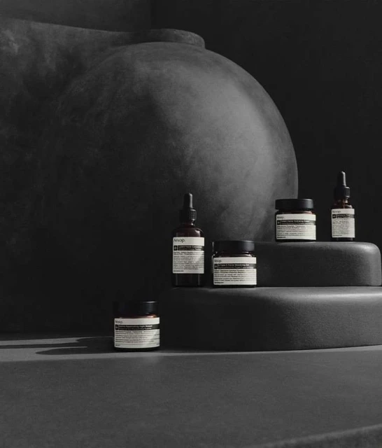 Several Aesop skincare products are arranged on tiered gray platforms against a dark backdrop with a dark, abstract sculpture.