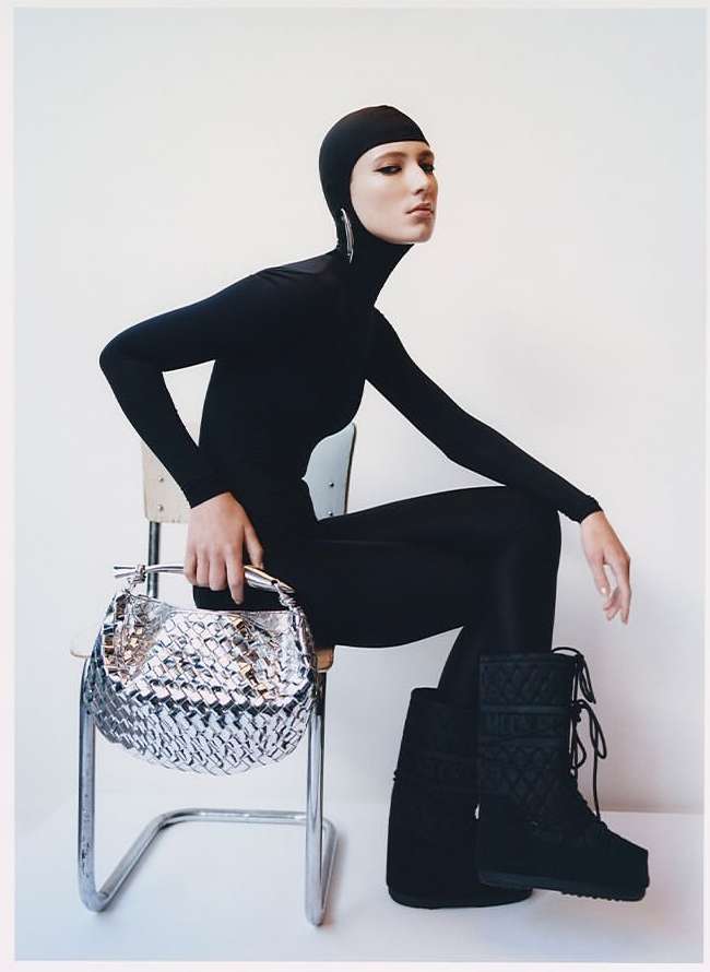 A fashion shot features a female model in a black bodysuit and boots, sitting on a chair and holding a silver handbag against a white backdrop.