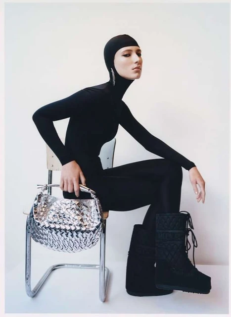 A fashion shot features a female model in a black bodysuit and boots, sitting on a chair and holding a silver handbag against a white backdrop.