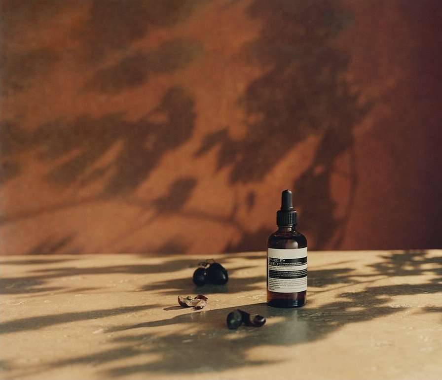 Still life with Aesop bottle, fruits, and foliage shadows on a beige surface against a textured orange-brown background.