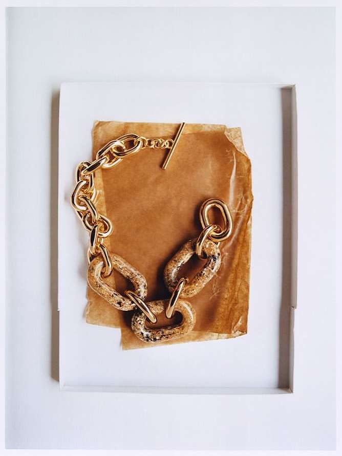 A close-up studio shot showcases a chunky gold chain bracelet with marbled links, displayed on a piece of brown paper against a white background.