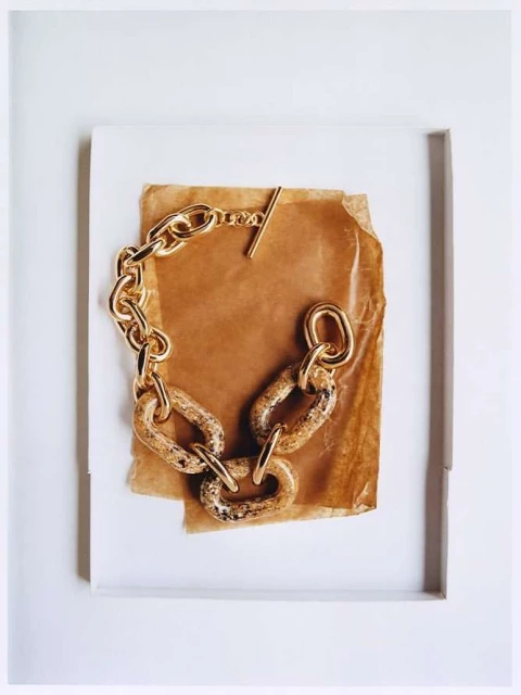 A close-up studio shot showcases a chunky gold chain bracelet with marbled links, displayed on a piece of brown paper against a white background.