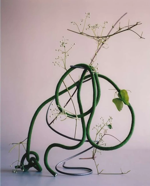 Still life of a green coiled rope atop a metal frame, with a dried floral arrangement against a soft, light pink backdrop.