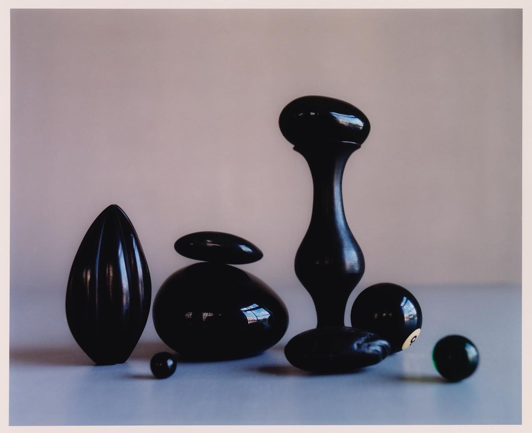 Still life photography features various black, glossy, abstract shapes and billiard balls on a light surface against a neutral background.