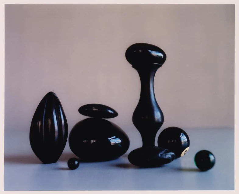 Still life photography features various black, glossy, abstract shapes and billiard balls on a light surface against a neutral background.