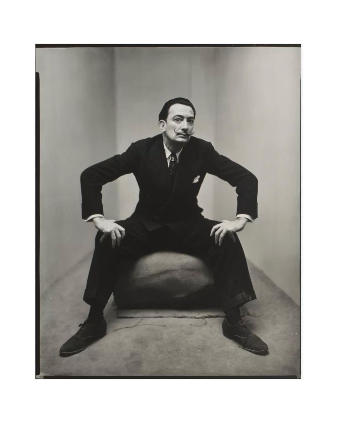 Monochrome portrait of a man in a suit sitting on a large rounded object with his hands on his knees and elbows out.
