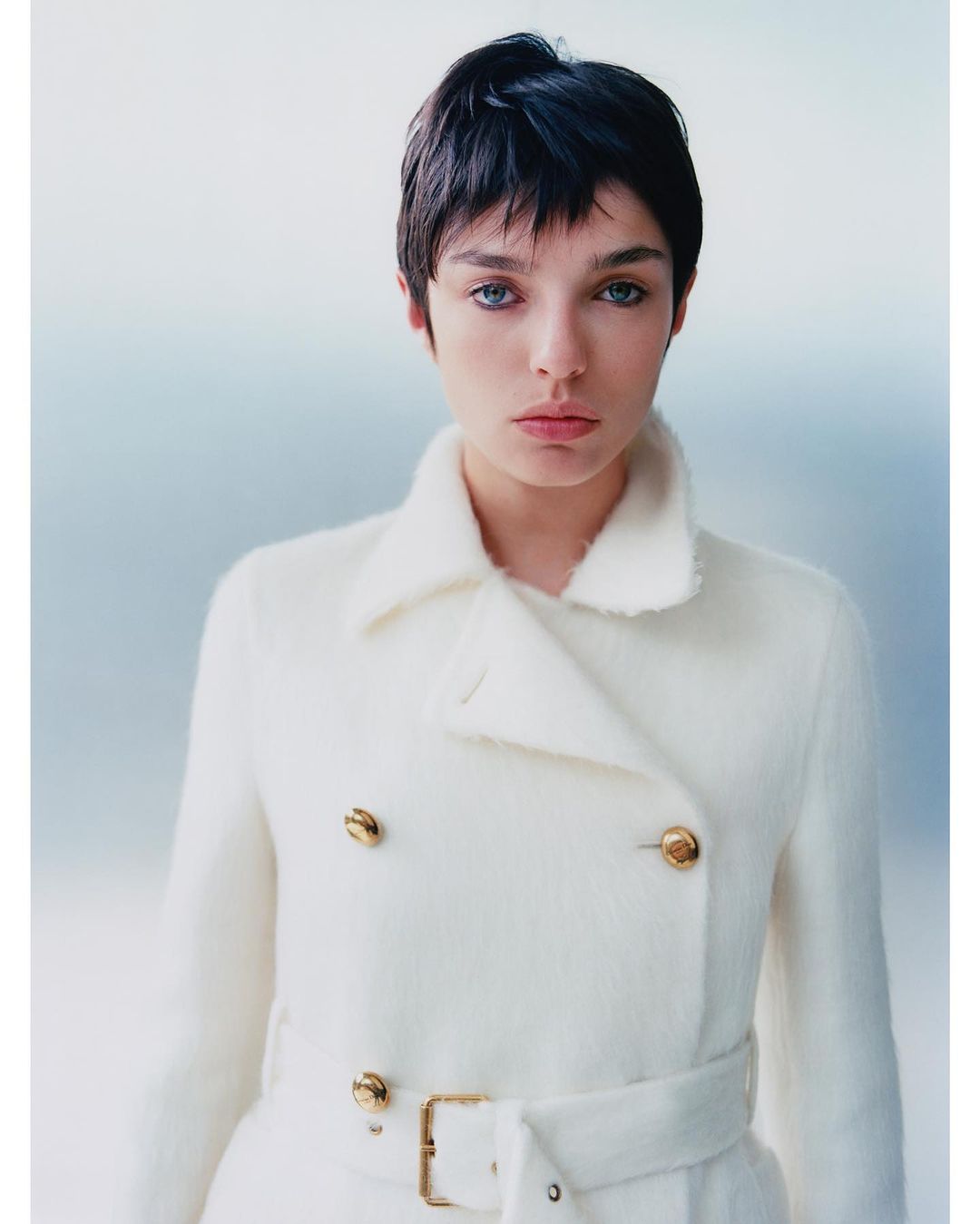 Close-up portrait of a young adult female with short, dark hair wearing a stylish white wool coat with gold buttons indoors.