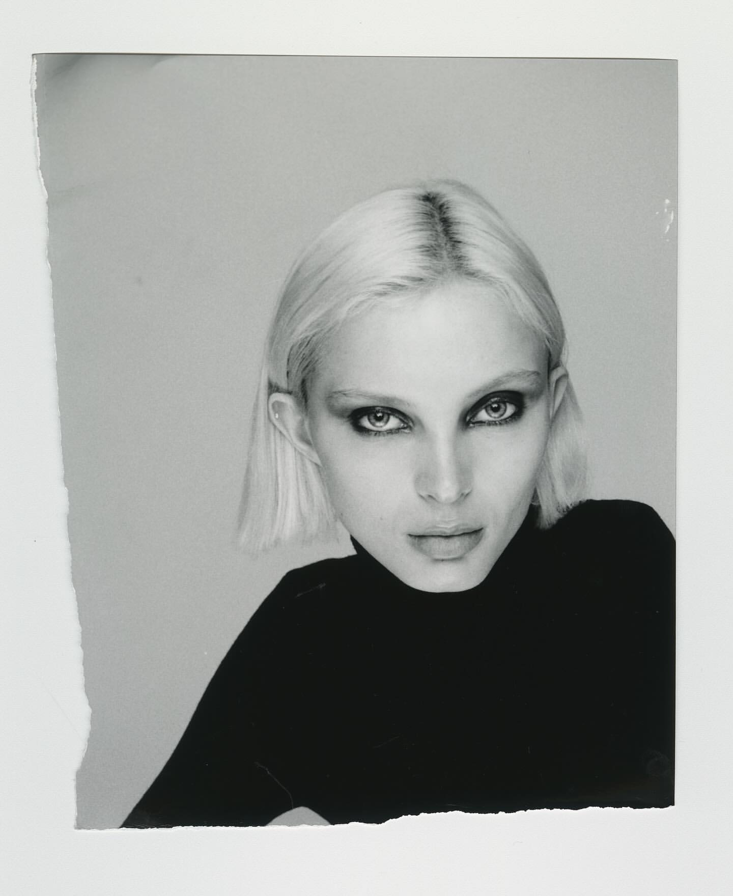 Monochrome close-up of a fair-skinned adult woman with short blonde hair, wearing a black top, and staring directly at the camera.