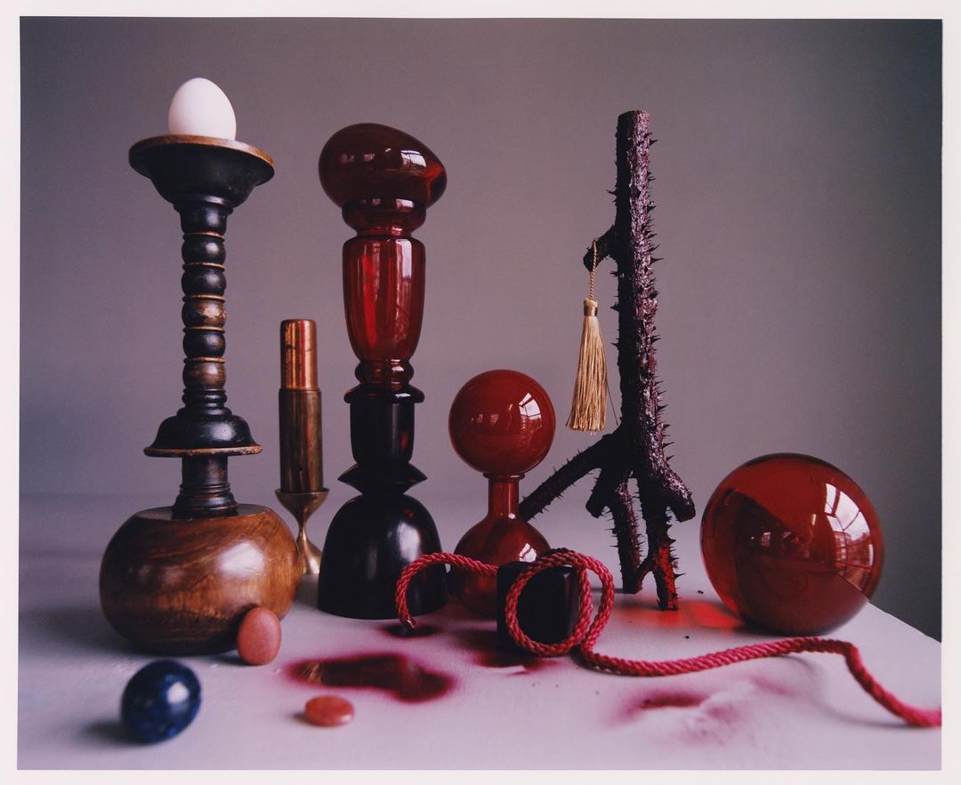 Still life photography featuring egg on wooden candlestick, red glass objects, branch, and red cord against a grey background.