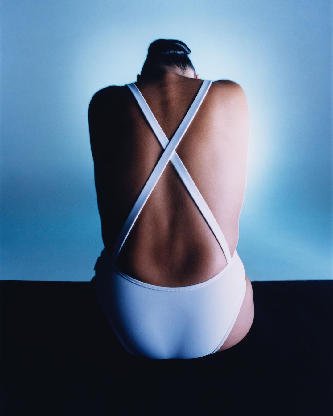 Back view of an adult woman with her hair up, wearing a white one-piece swimsuit with crisscross straps against a blue gradient background.