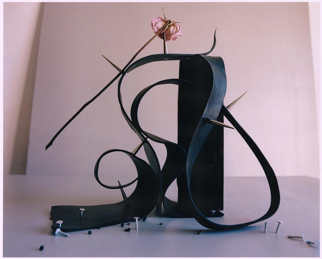 Abstract still life: black leather strips arranged in sculpture with nails, thorns, and a dried rose on a light grey table.