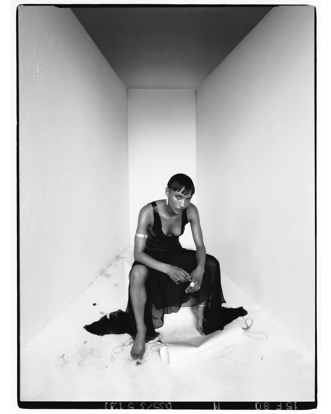 Monochrome studio portrait of a woman in a dark dress, seated in a white box. Ethereal lighting highlights her expression and bare feet.