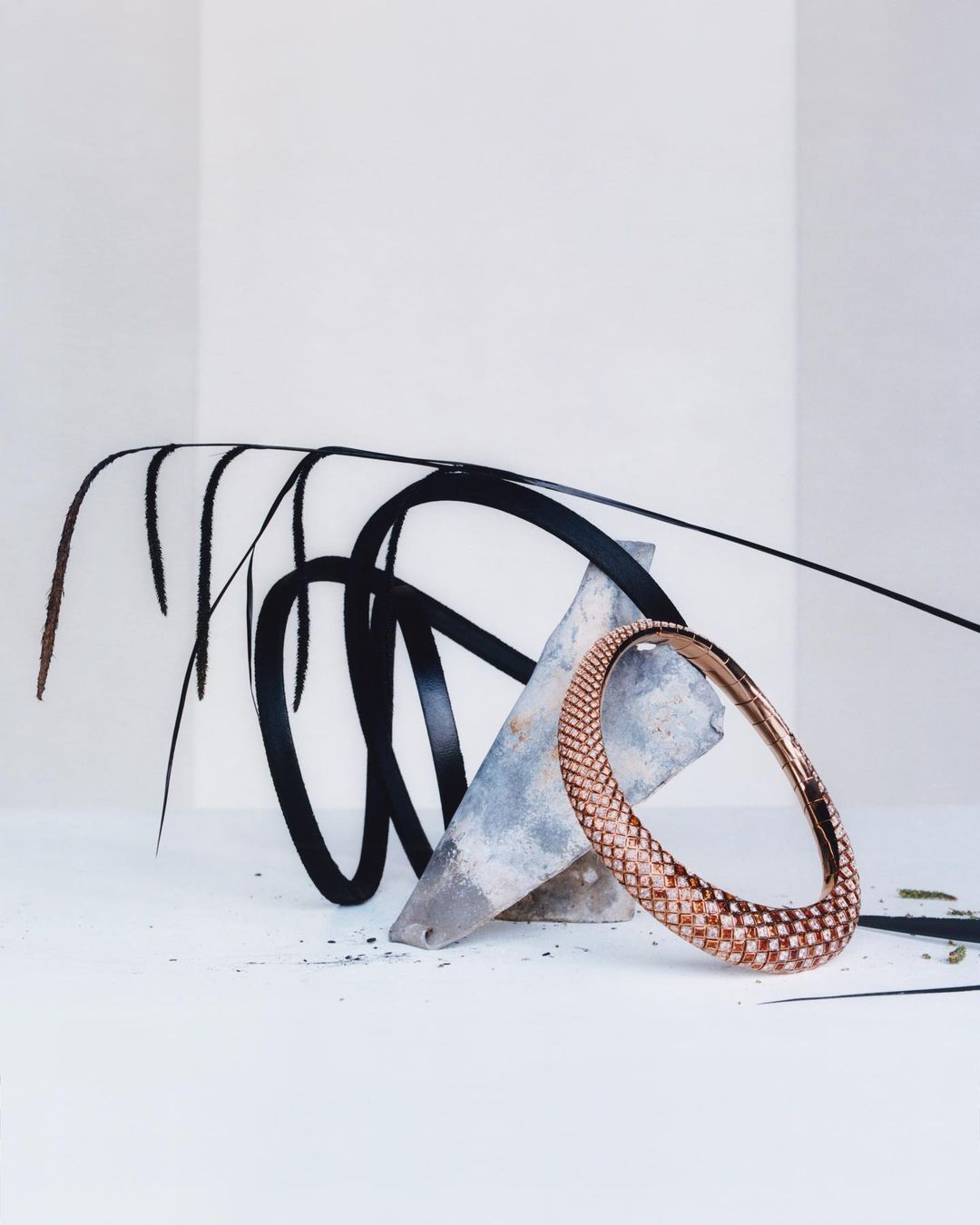 Studio shot of a rose gold bangle with a woven texture, contrasted against abstract shapes and shadows on a white backdrop.