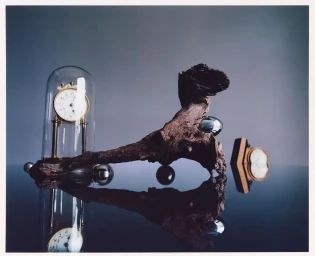 Still life photography by David Abrahams featuring a weathered piece of wood, a clock under a glass dome, and polished metallic spheres on a reflective surface.