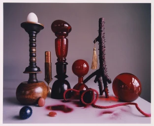Still life arrangement of sculptural objects including red glass, wooden candlesticks, and a white egg on a table
