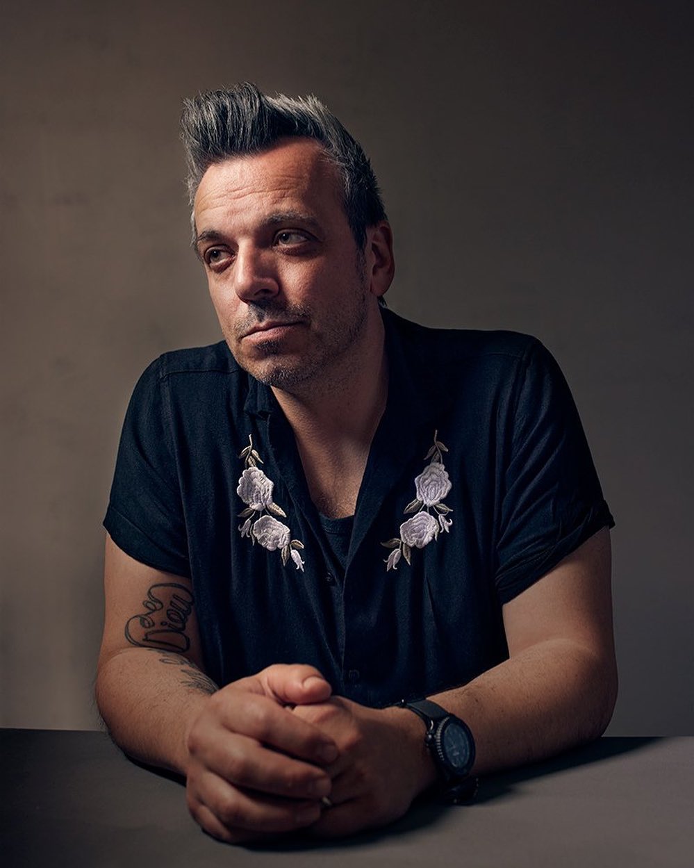 Portrait of an adult male with dark hair with gray streaks wearing a floral-embroidered shirt with folded hands, shot in a photo studio.