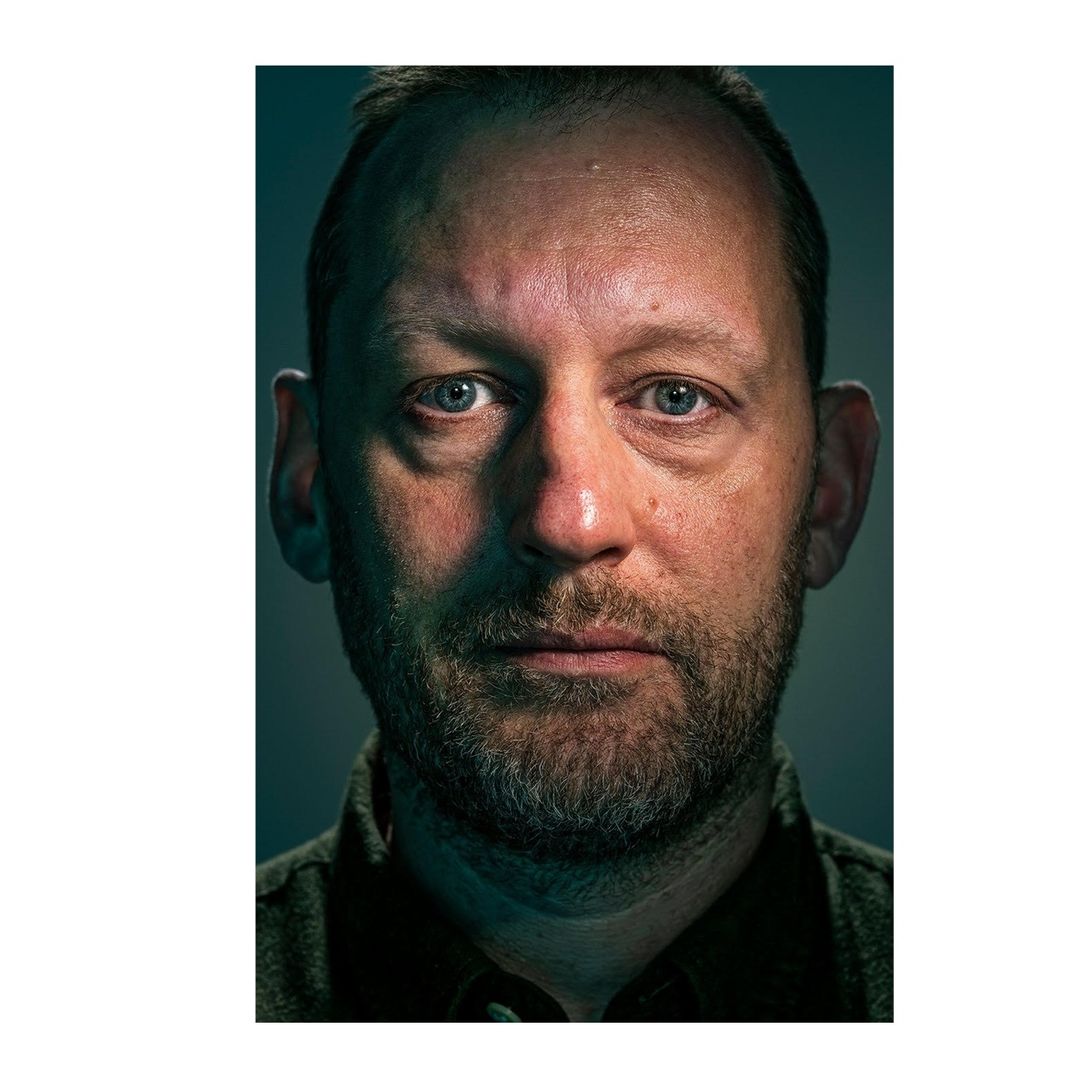 Close-up portrait of an adult male with a beard, looking directly at the camera with a neutral expression, captured in a studio setting.