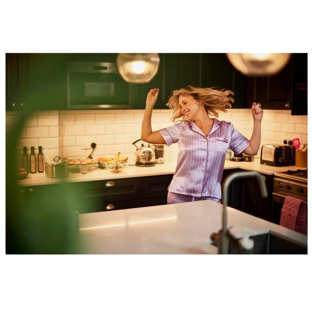 Woman in pajamas dances happily in a brightly lit kitchen with modern appliances and warm, inviting lighting in a cozy home interior.