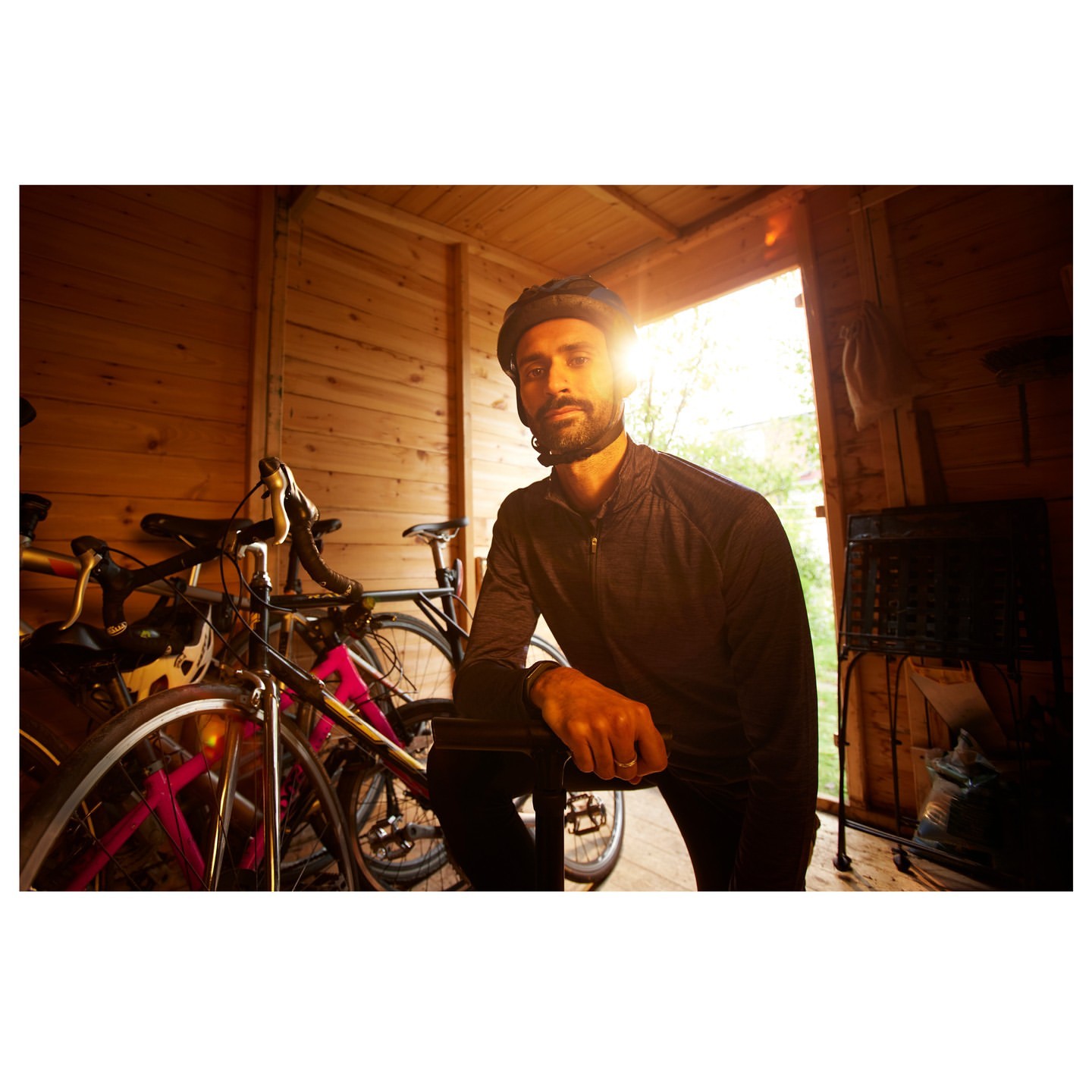 Adult male cyclist in helmet and athletic wear kneeling indoors in a rustic wooden shed with several bikes, backlit by bright sunlight.