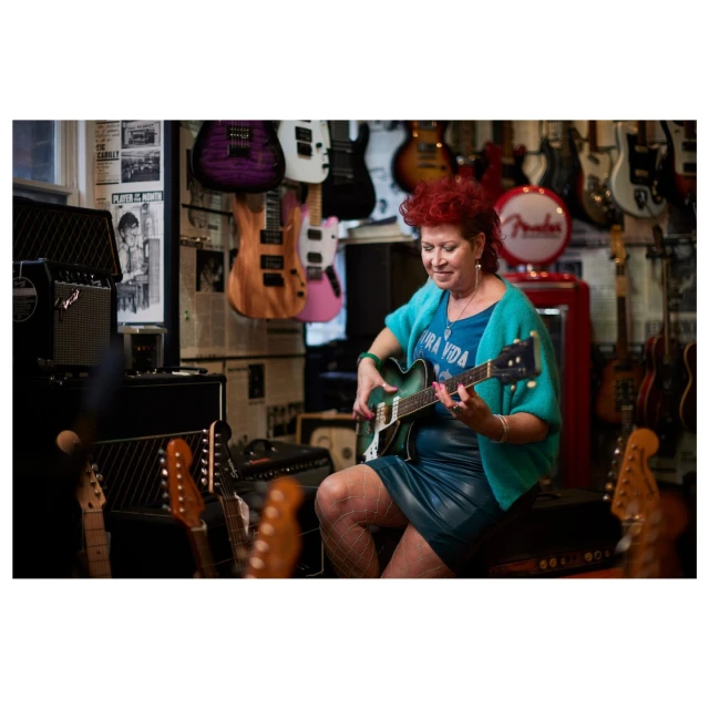 Woman with red hair plays a green electric guitar in a music store surrounded by guitars and amps, showcasing her musical talent.