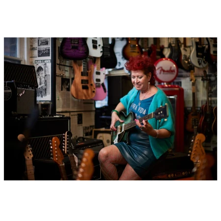 Woman with red hair plays a green electric guitar in a music store surrounded by guitars and amps, showcasing her musical talent.