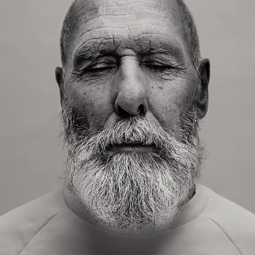 Monochrome portrait of an elderly man with a gray beard and closed eyes against a light gray background.