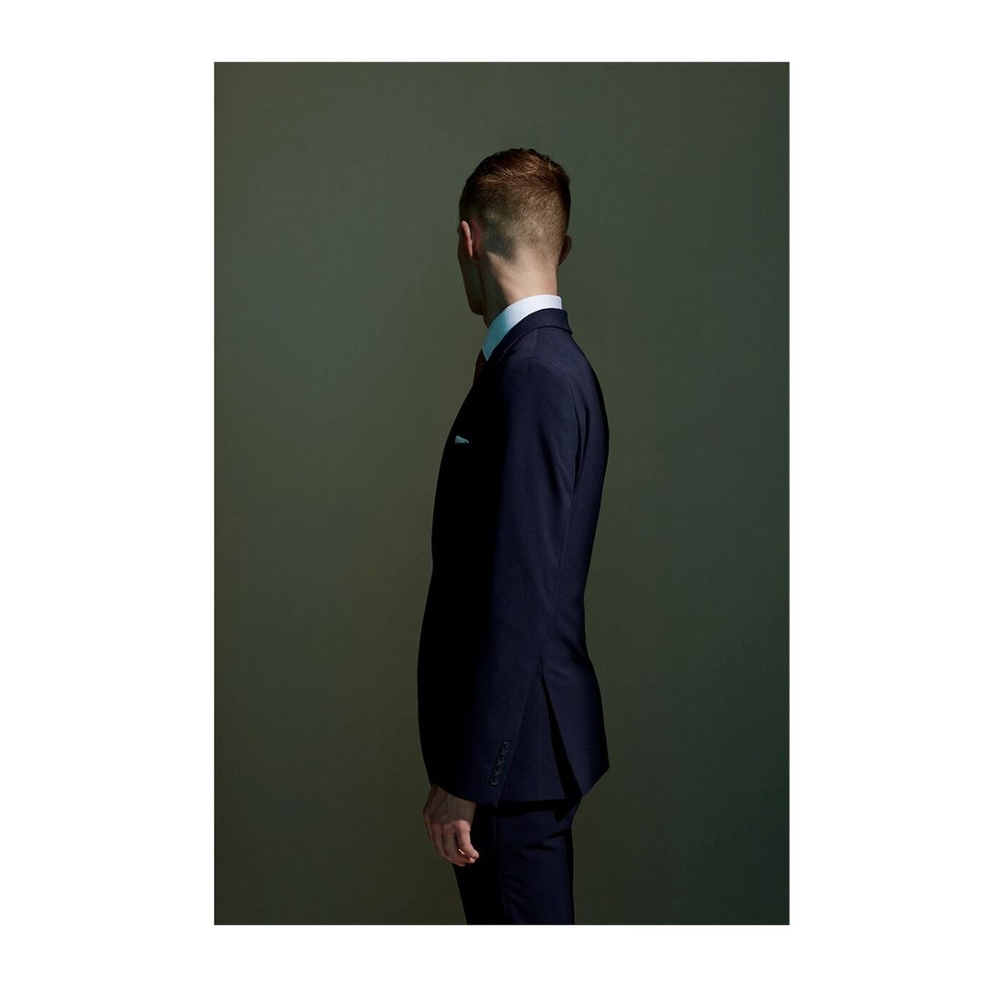 An adult male is shown from the side in a dark blue suit and light blue shirt against a neutral background in a studio setting.