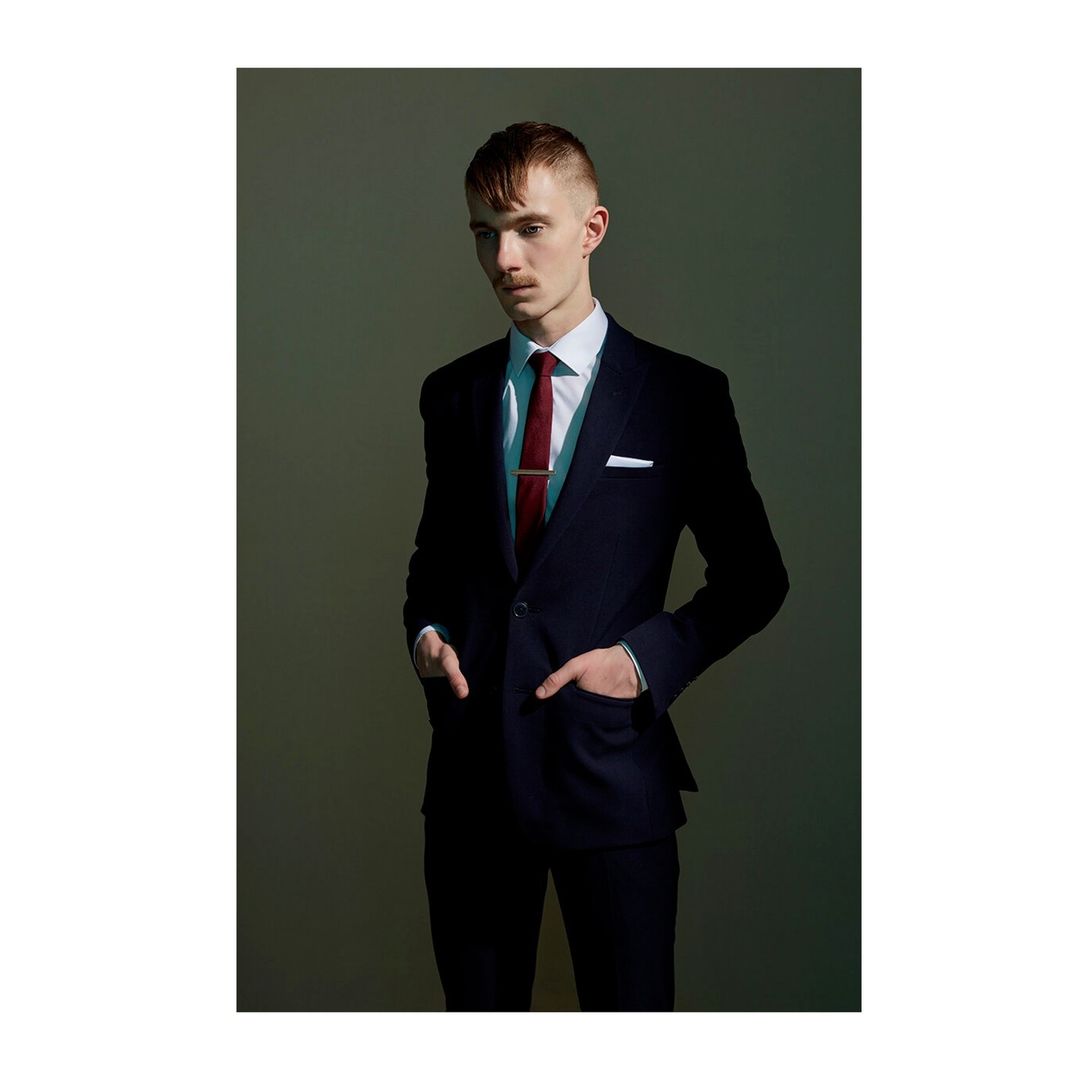 Portrait of a young adult man in a dark suit, white shirt, and burgundy tie standing in a studio with his hands in his pockets.