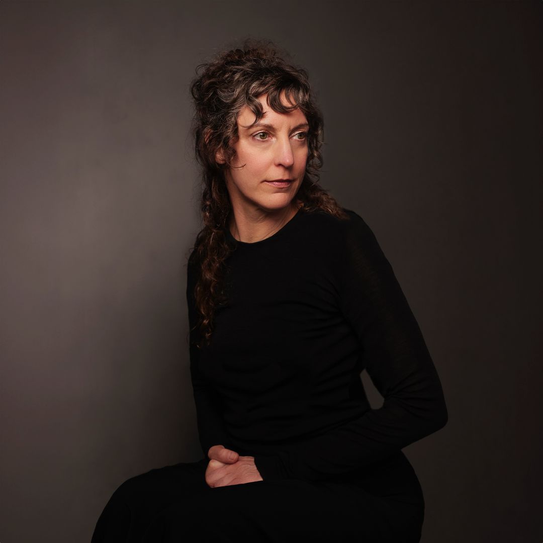 Portrait of a woman with curly brown hair wearing a long-sleeved black top against a dark grey background by David Boni