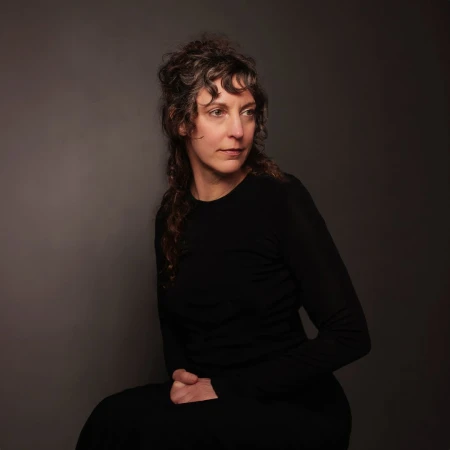 Portrait of a woman with curly brown hair wearing a long-sleeved black top against a dark grey background by David Boni
