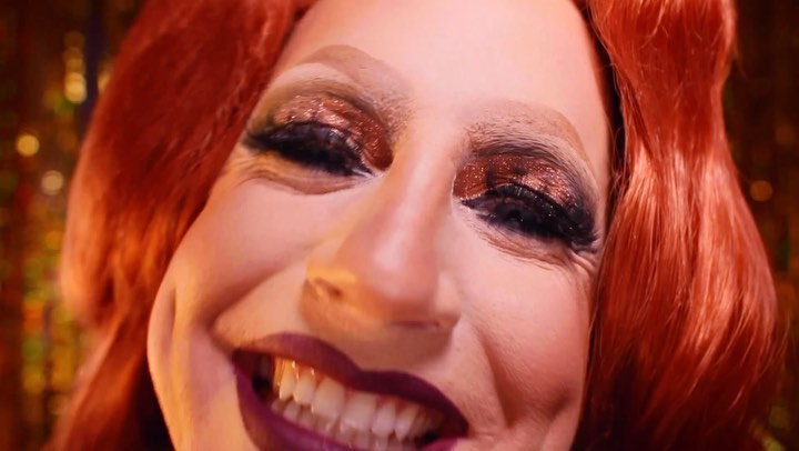 Close-up studio portrait of a smiling redhead adult wearing gold eyeshadow and dark lipstick, set against a blurred gold background.