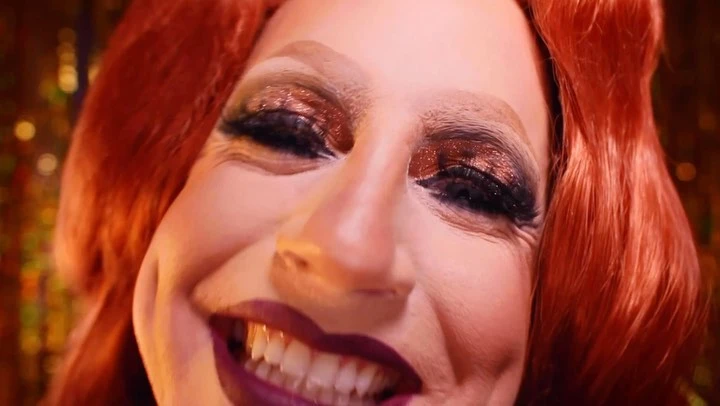 Close-up studio portrait of a smiling redhead adult wearing gold eyeshadow and dark lipstick, set against a blurred gold background.