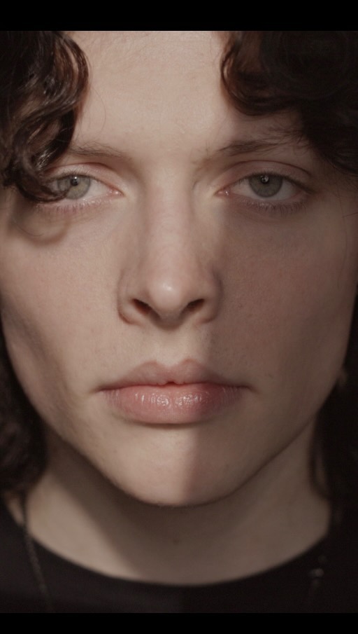 Close-up portrait of a young person with dark curly hair and light eyes, illuminated by a vertical strip of light across the face.
