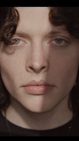 Close-up portrait of a young person with dark curly hair and light eyes, illuminated by a vertical strip of light across the face.